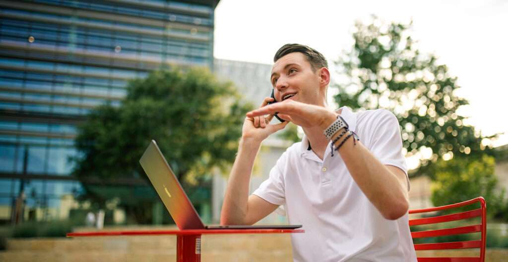 Man sitting at outdoor table with laptop while on the phone
